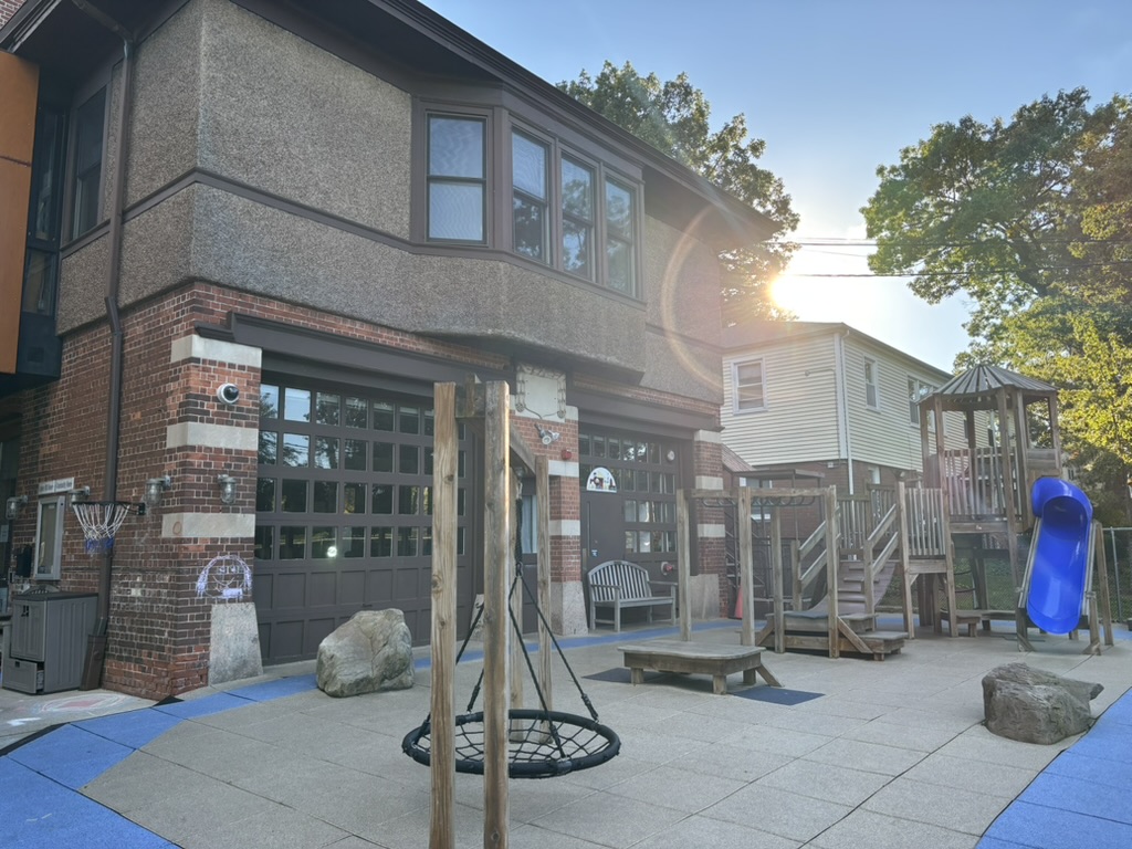 Closer view of Calvin Hill Daycare Center frontal playground apparatuses, featuring a spider-web swing, monkey bars, a basketball hoop, space for chalk drawings, and a larger climbing structure with a slide and bridge. Original firehouse garage doors are also in view. 