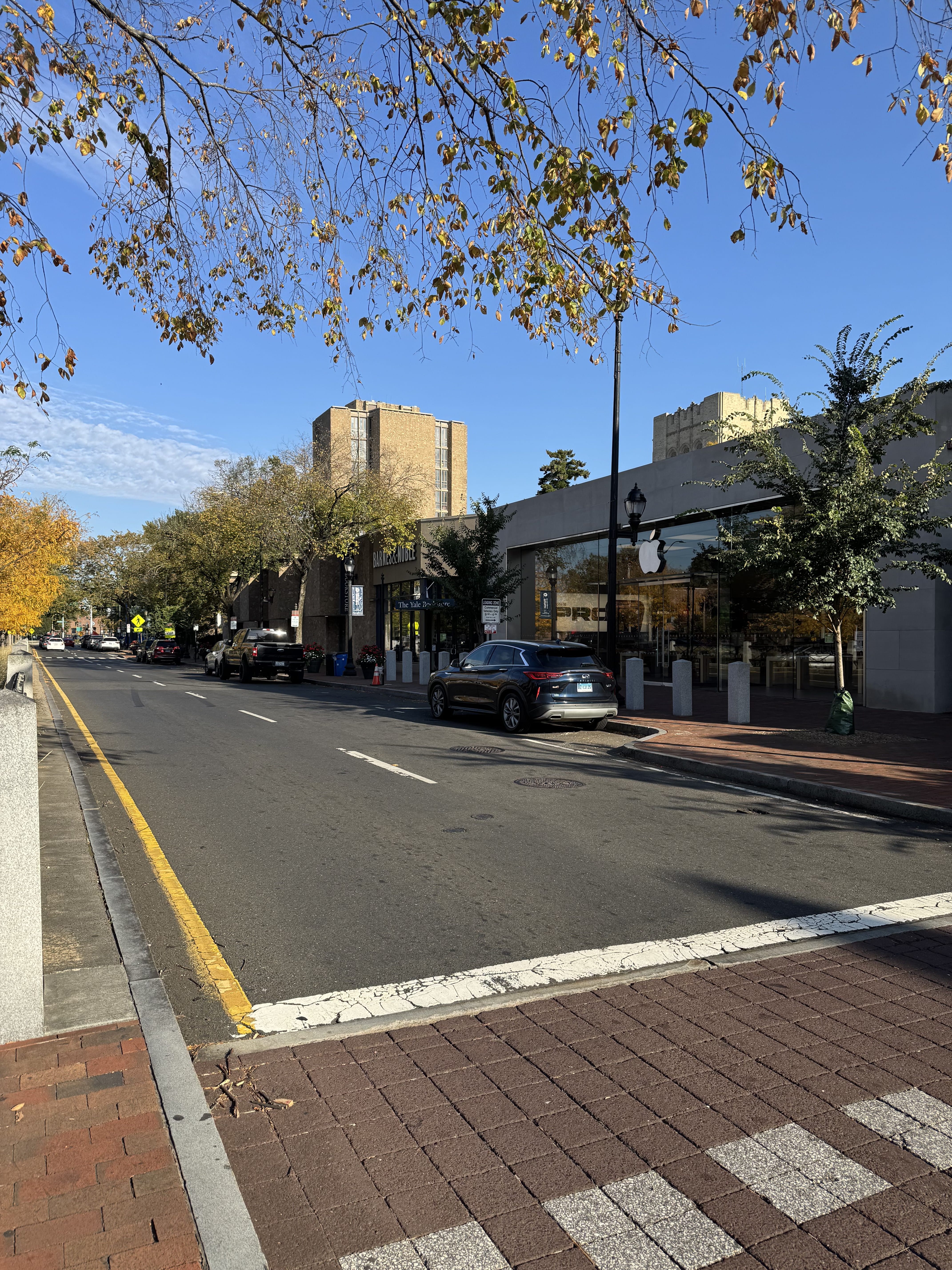 :  Looking up Broadway past the Apple store towards the Yale Bookstore. The street has brick sidewalks, granite bollard, and trees lining it. The campus buildings rise behind the shops.
