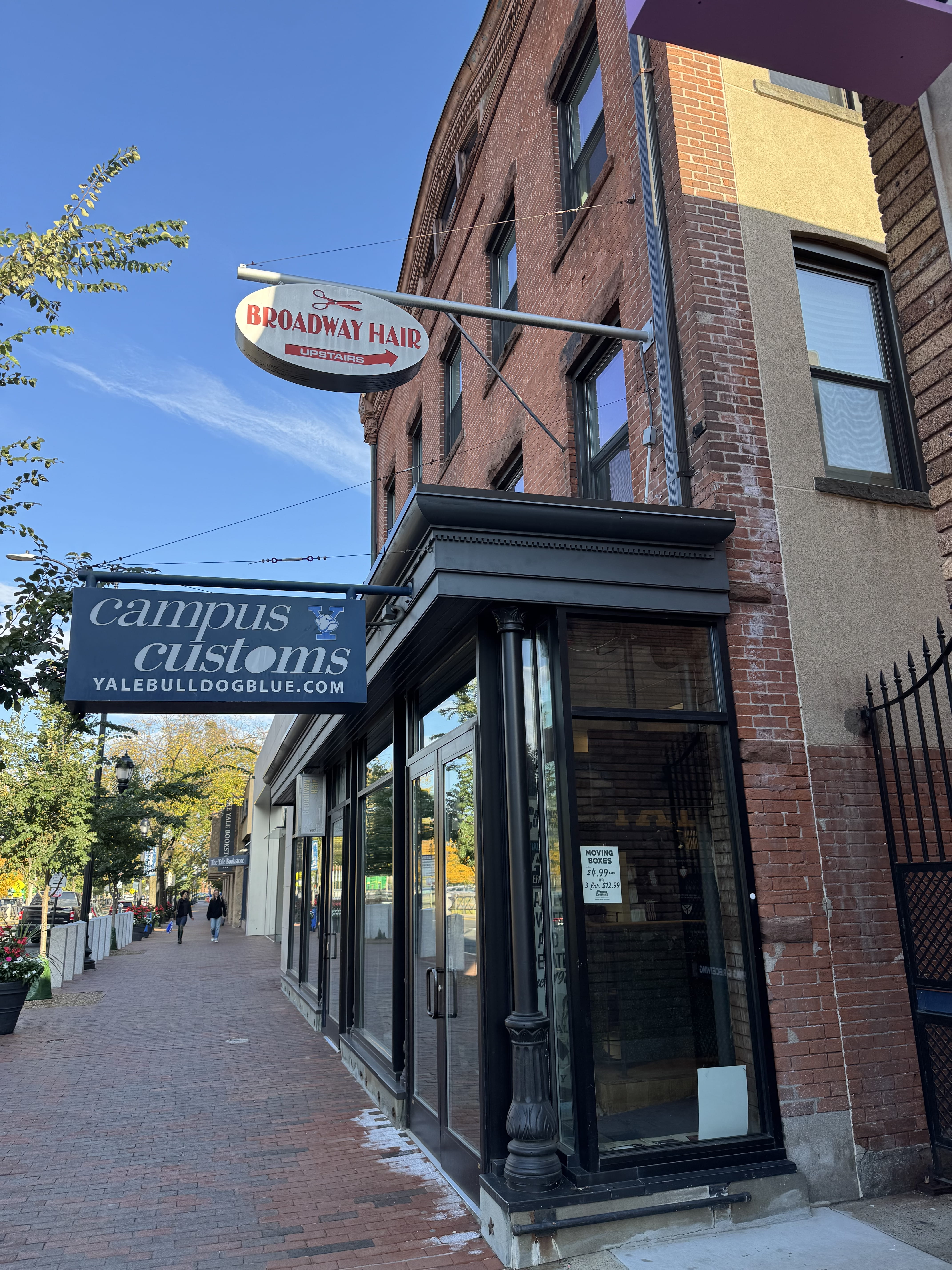 Angled view of the Campus Customs storefront from the sidewalk. A dark metal column marks the corner of the ground-floor entrance, with the brick upper floors rising above it.