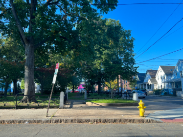 This street view of the public Trowbridge Square and Cedar Street shows a small slice of the neighborhood surrounding 167 Cedar St. On the righthand side, we notice a row of Queen-Anne style/Colonial Revival-style houses, likely built pre-1900. The park has a low fence of wrought iron and stone, with an open space welcoming visitors in.
