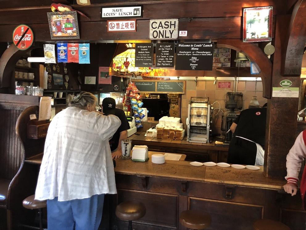 View of Interior Counter of Louis’ Lunch. Parts of the counter are the original, from the building prior to its move. This photograph also shows some of the building’s hung memorabilia, including the “Save Louis’ Lunch” sign and the No Coca-Cola sign, a gesture to the Lassen’ longtime partnership in selling New Haven-based Foxon Park Beverages. The photograph also includes etched signatures in the counter and building’s woodwork, some of which date back to the early twentieth century.